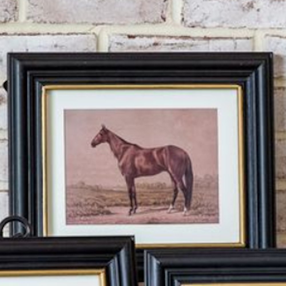 Framed brown horse print in a black frame with gold inner trim against a brick wall.