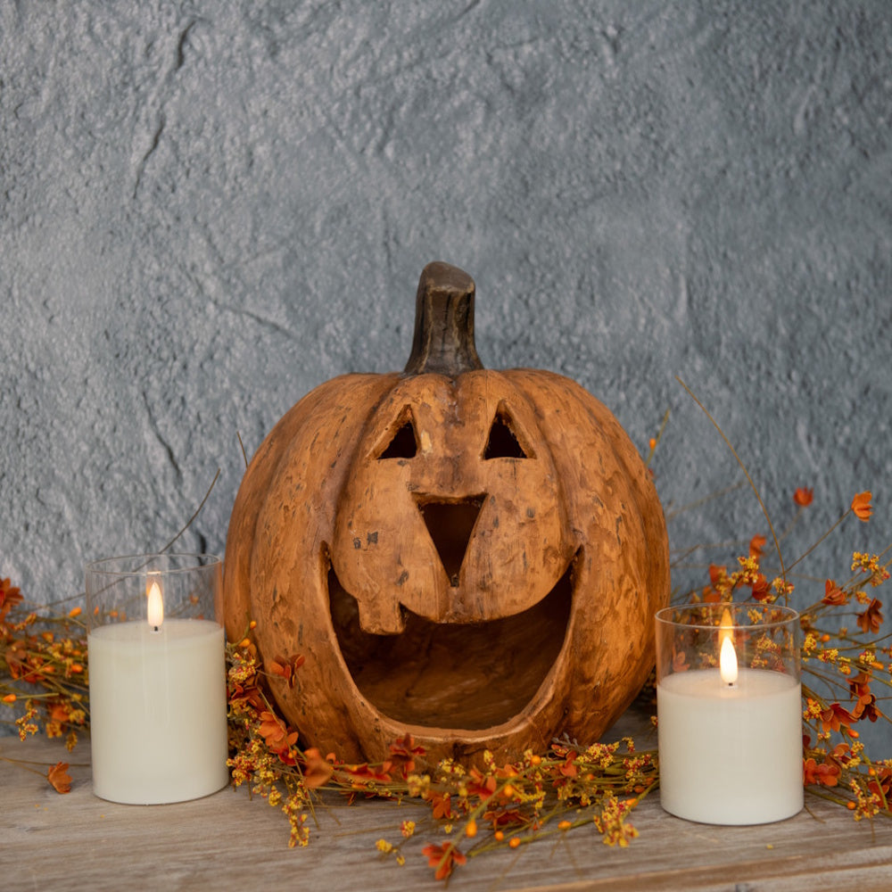 Carved terracotta jack-o'-lantern flanked by lit glass candles and autumn foliage against a grey wall.