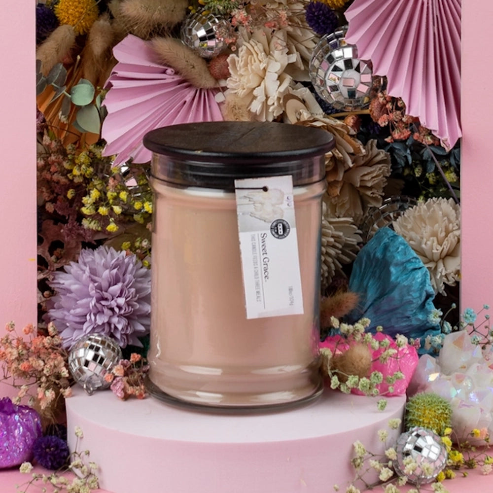 Sweet Grace large glass jar candle displayed on a pedestal surrounded by colorful dried flowers.