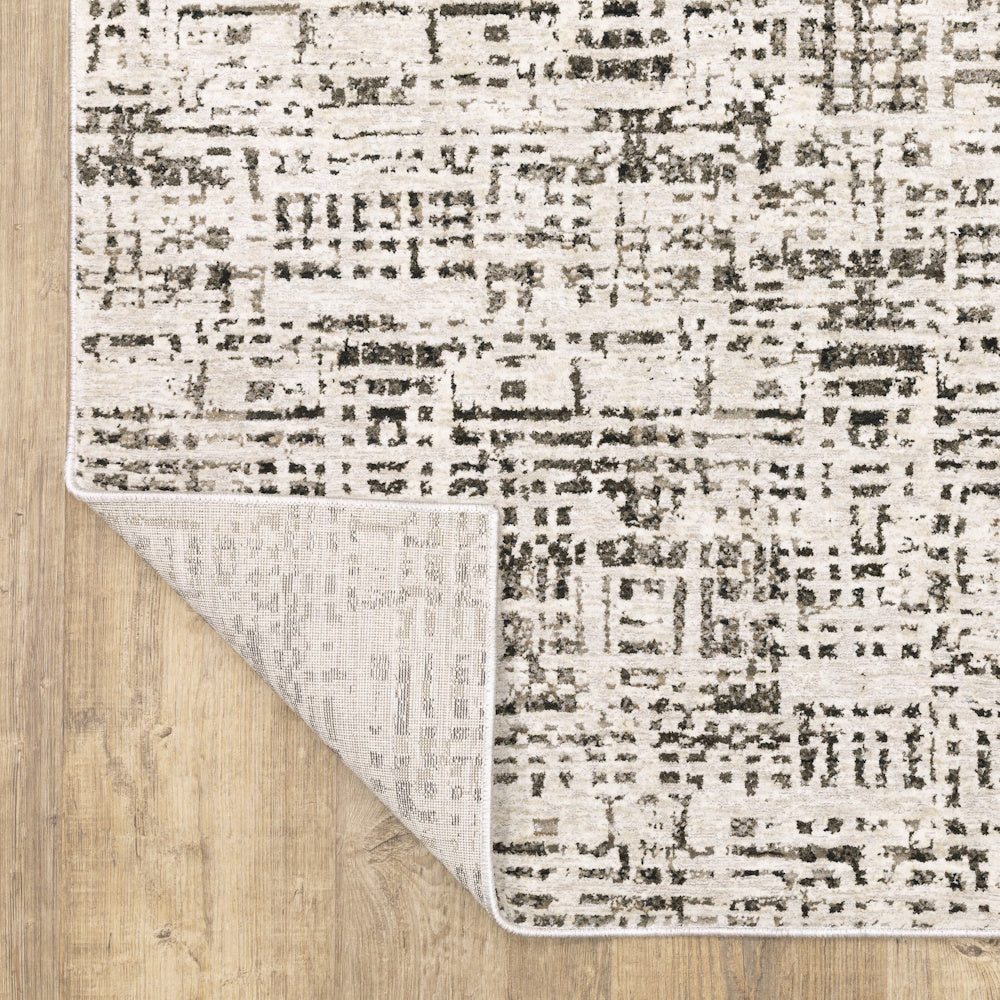 Corner of the gray and cream area rug folded back to show the backing material on a wooden floor.