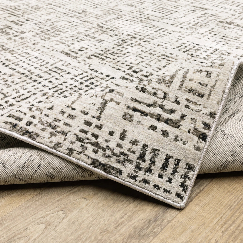 Corner of the gray and cream area rug folded to show its finished binding edge and backing on a wooden floor.
