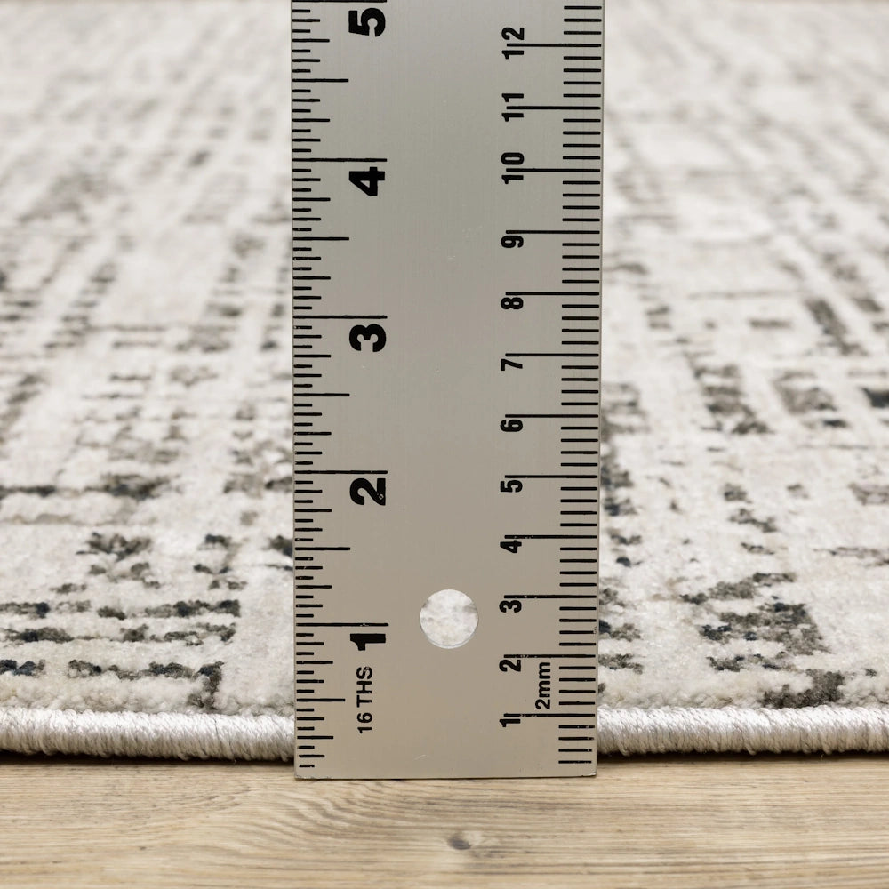 Close-up view of the gray and cream area rug with a ruler placed vertically next to it to show its thickness on a wooden floor.