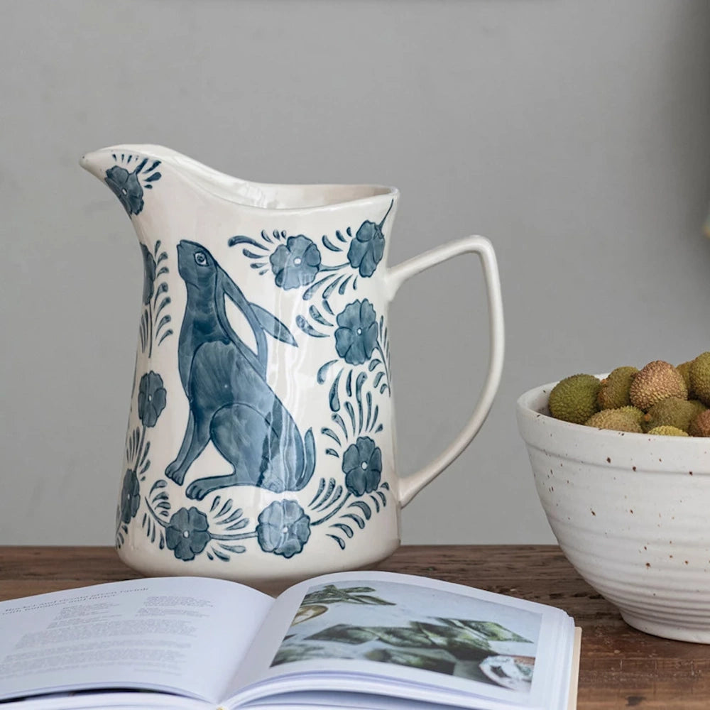 Hand-painted blue and white stoneware pitcher with a rabbit and floral design next to a bowl of fruit.