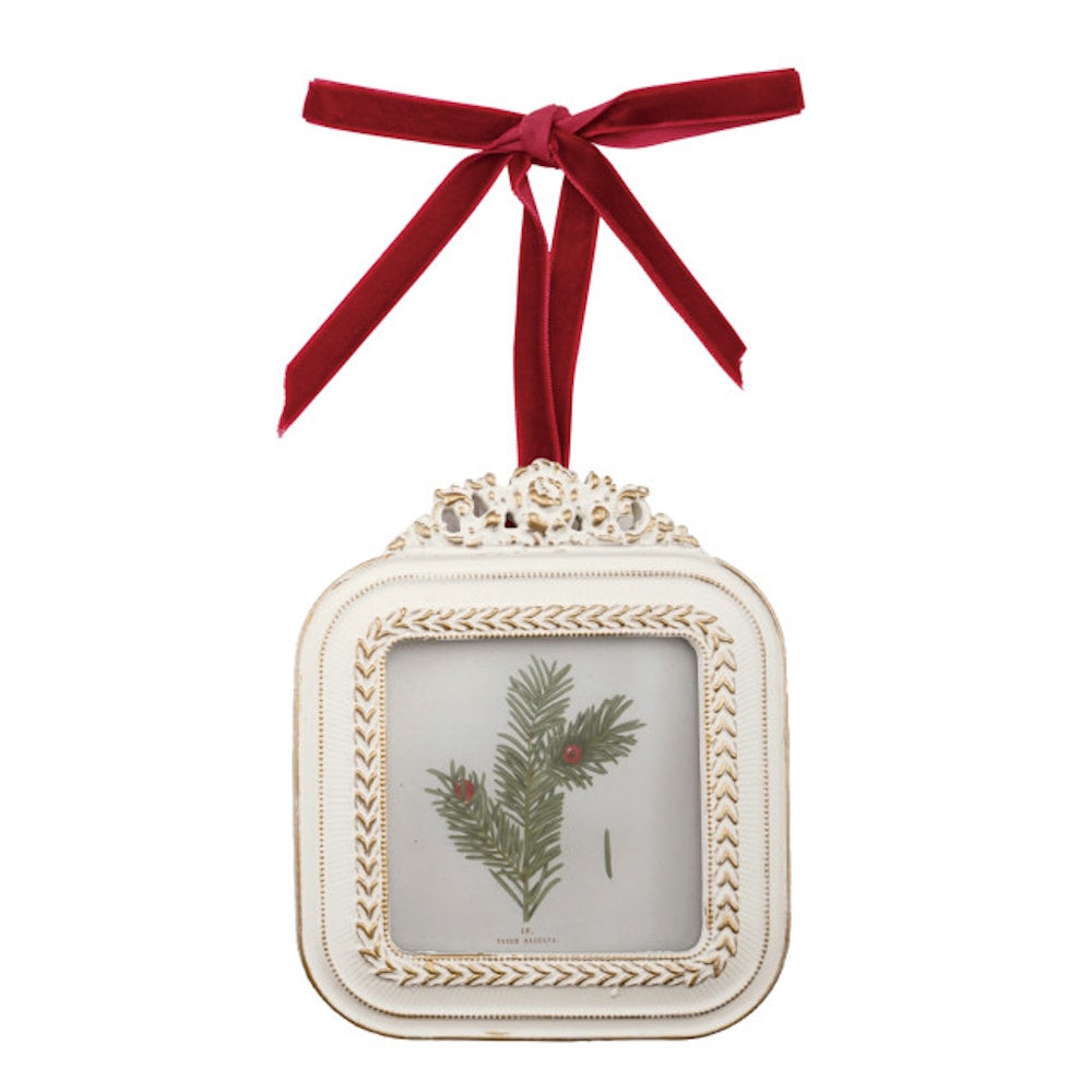 An ornate white and gold picture frame ornament with a botanical pine print and red velvet ribbon.
