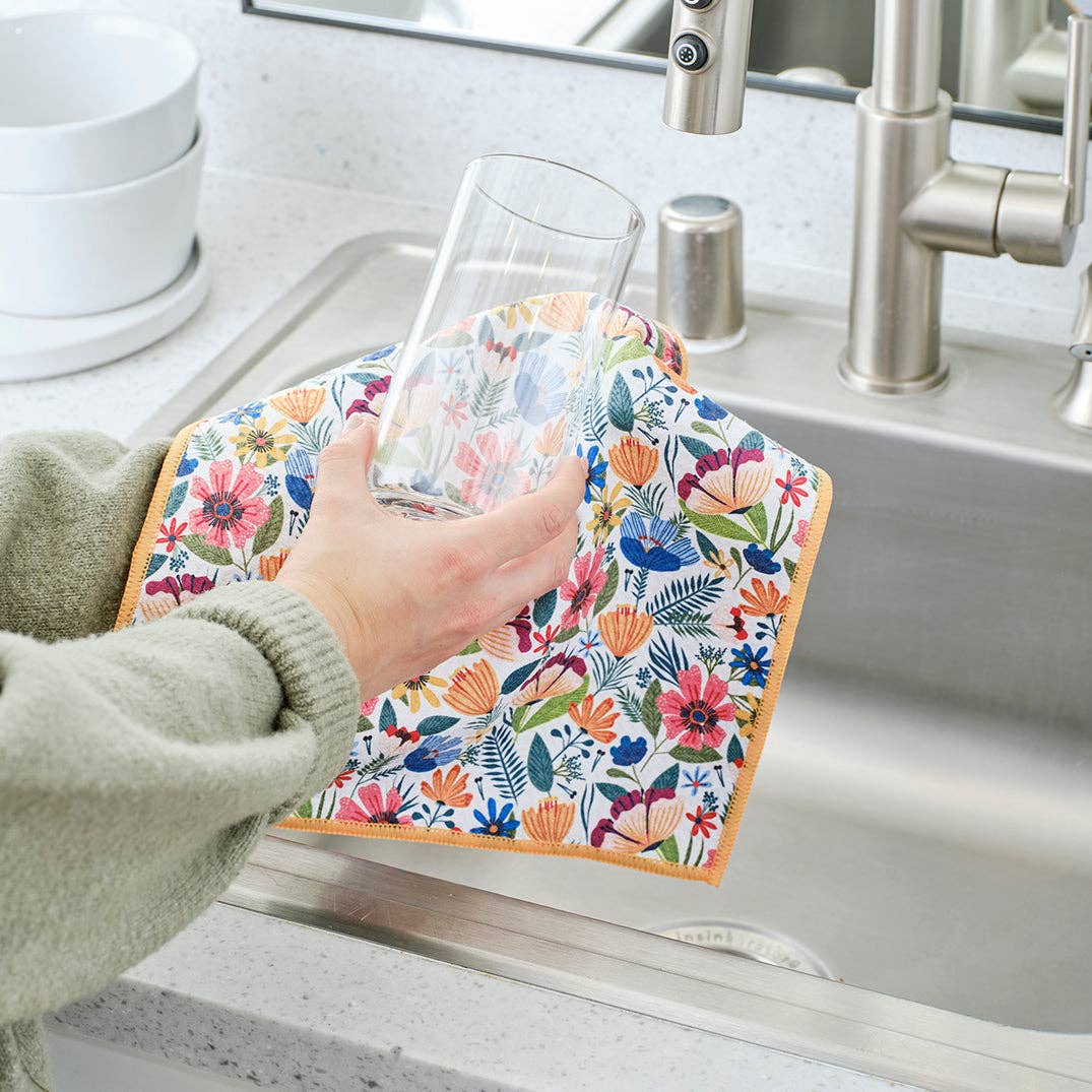 Person using a floral patterned dishcloth to dry a glass in a kitchen sink.