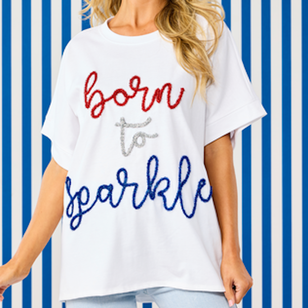 White oversized t-shirt with "born to sparkle" in red, silver, and blue sequins against a blue striped background.