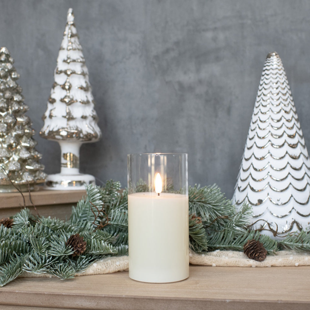 White flameless candle glowing in a glass cylinder surrounded by pine garland and decorative Christmas trees.
