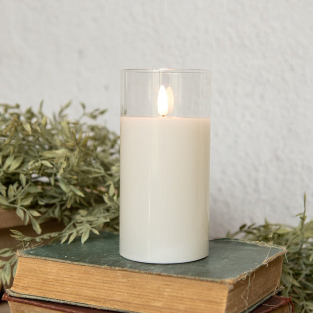 White pillar candle with realistic flame inside a clear glass cylinder, resting on vintage books.
