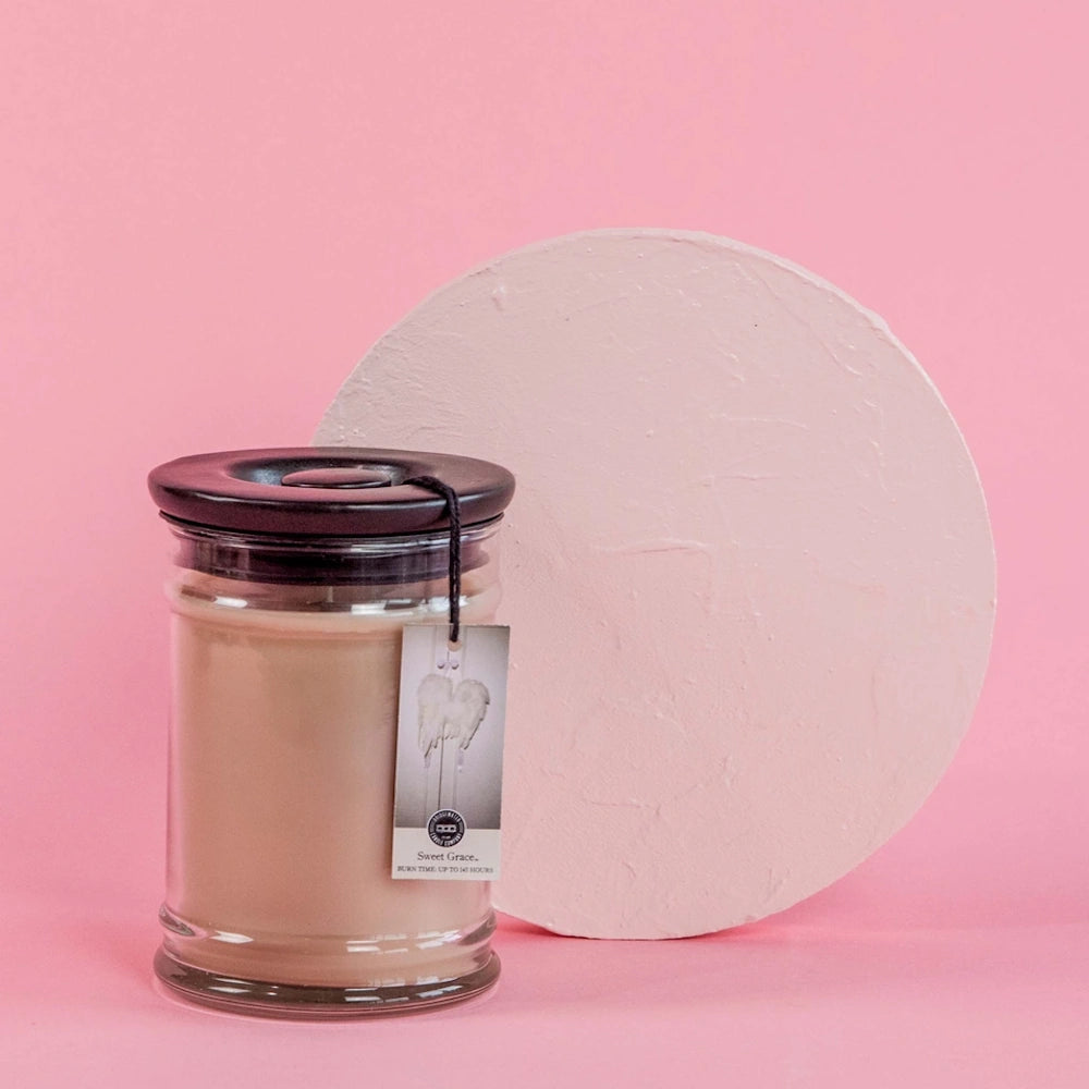 Sweet Grace jar candle with dark lid and hangtag in front of a textured pink backdrop.