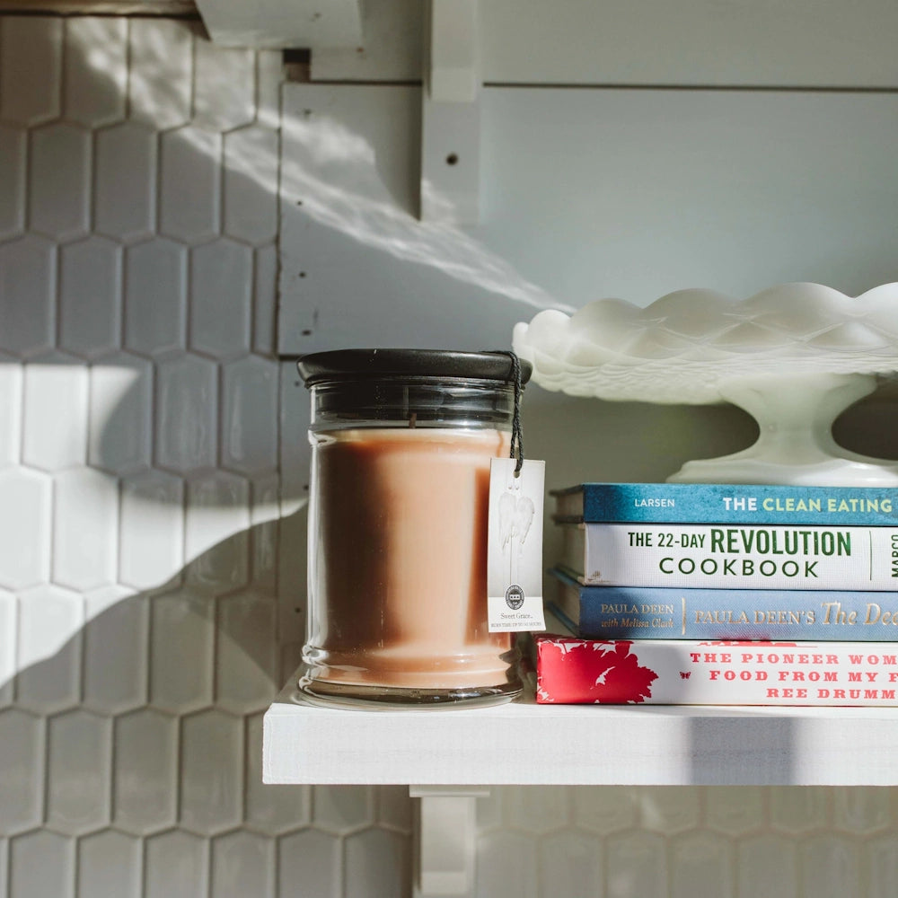 Sweet Grace glass jar candle with a black lid resting next to stacked cookbooks on a white shelf.