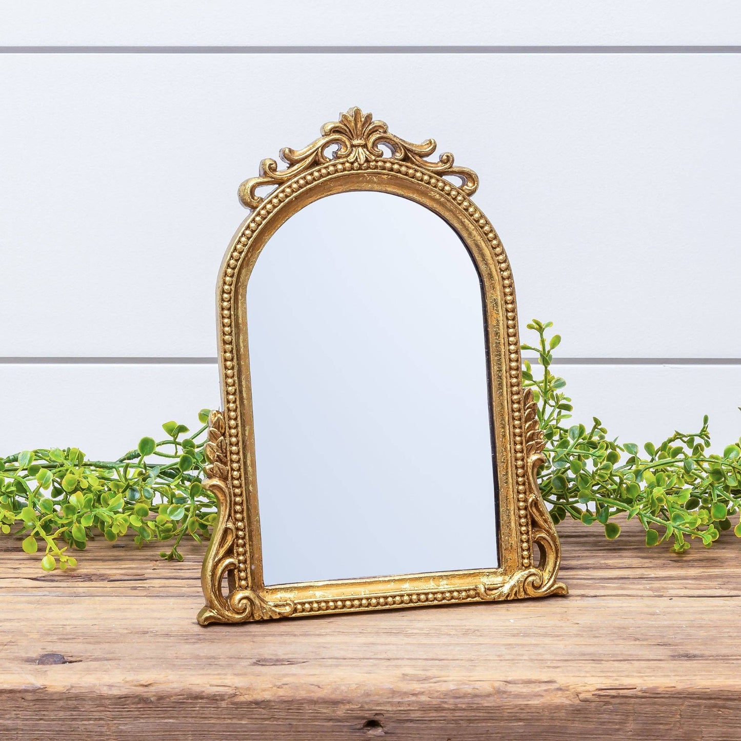 Arched gold leaf tabletop mirror with ornate beaded frame and easel back on a rustic wooden surface with greenery.