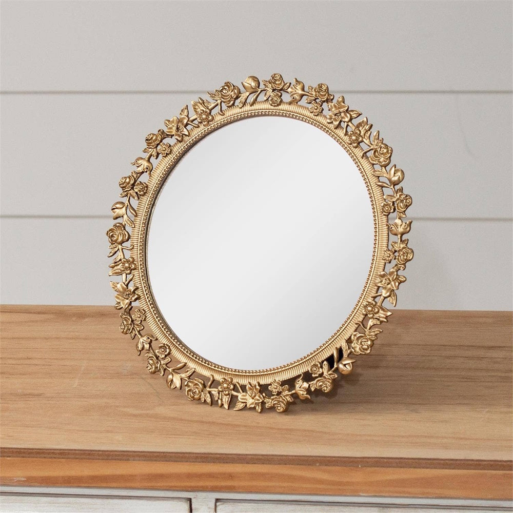 Antique-style round gold table mirror with an ornate floral rose frame on a wooden surface.
