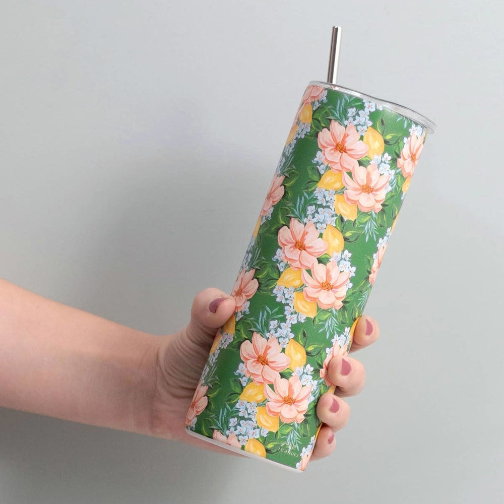 Hand holding a tall, green tumbler patterned with pink flowers, lemons, and a metal straw against a gray wall.