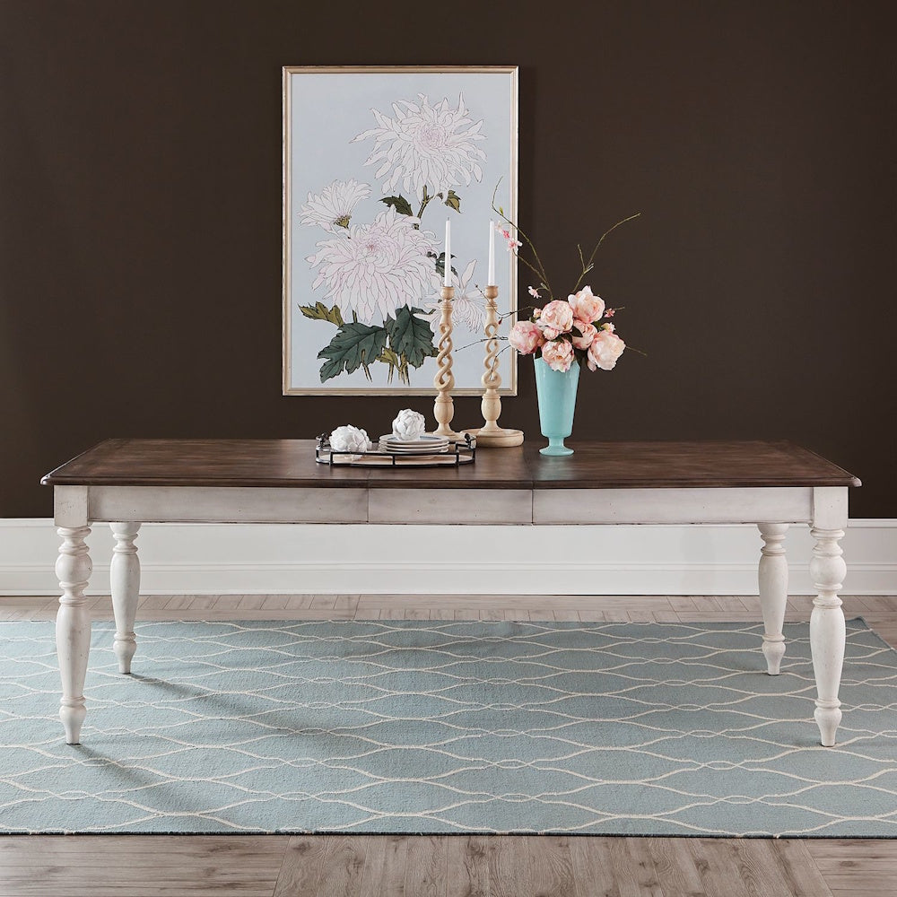 Wooden dining table with white legs on a blue patterned rug, decorated with flowers and artwork.