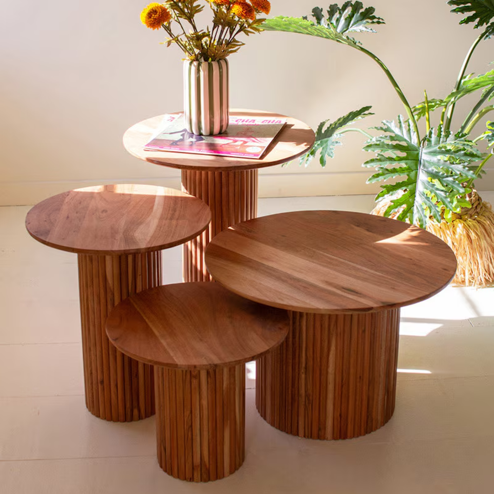 Four round acacia wood accent tables with ribbed pedestal bases styled together in a sunlit room.