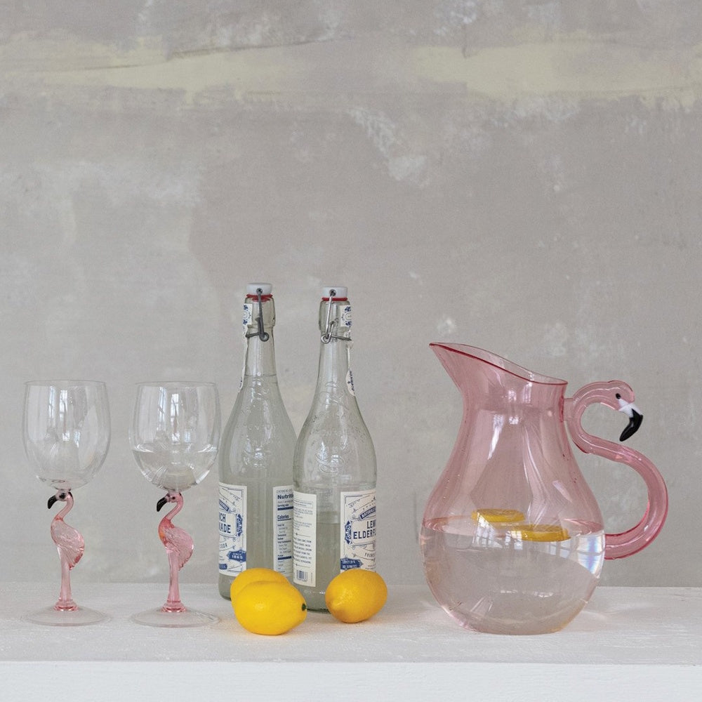 Pink flamingo pitcher and matching wine glasses on a table next to glass bottles and fresh lemons.