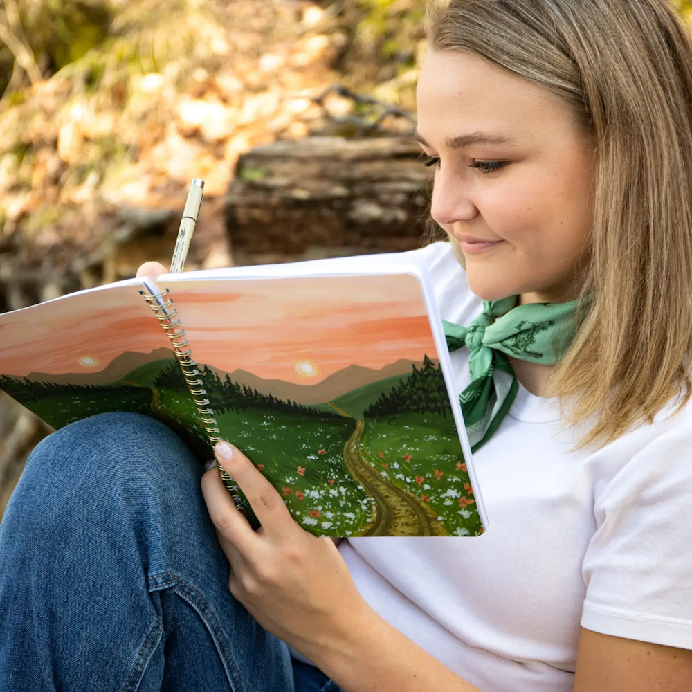 A woman smiling while holding a pen and looking at a notebook featuring a painted landscape cover.