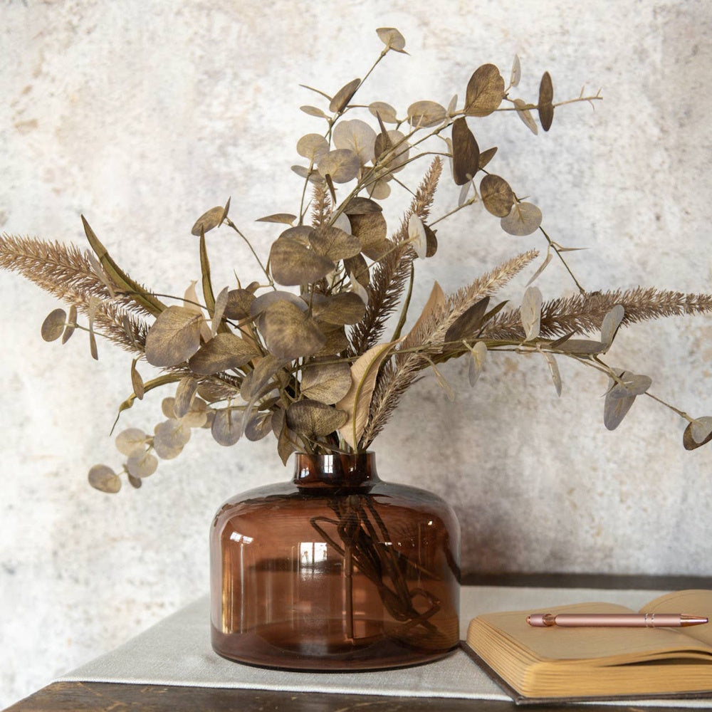 Amber glass vase with dried eucalyptus branches next to an open notebook on a table.