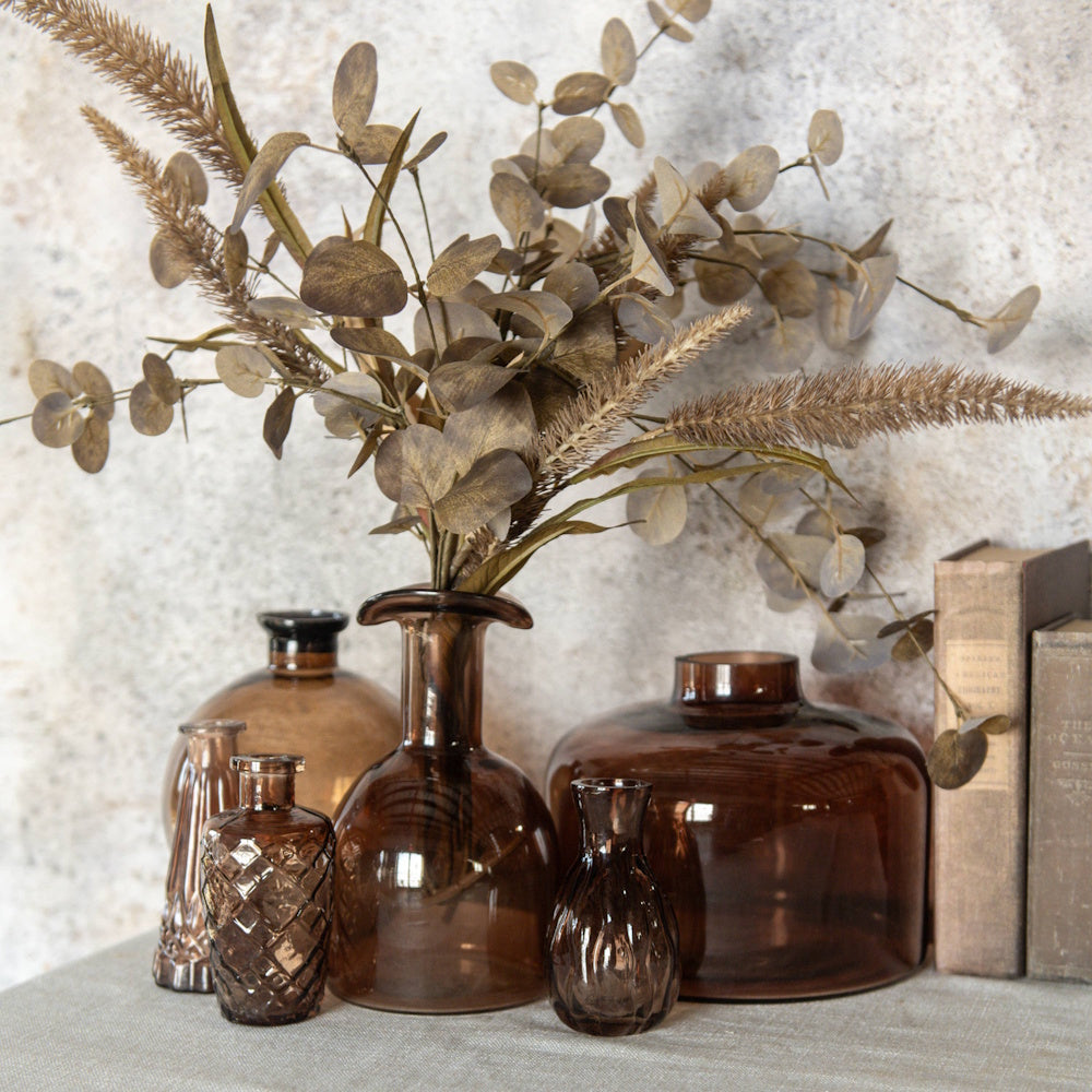 Assorted amber glass vases with dried foliage and books against a rustic wall.