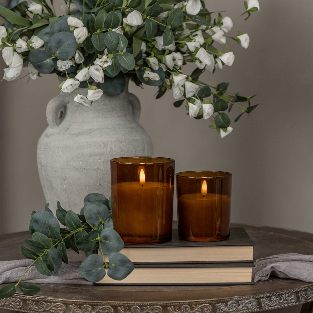 Two glowing amber glass candles on stacked books beside green eucalyptus and white floral arrangements.