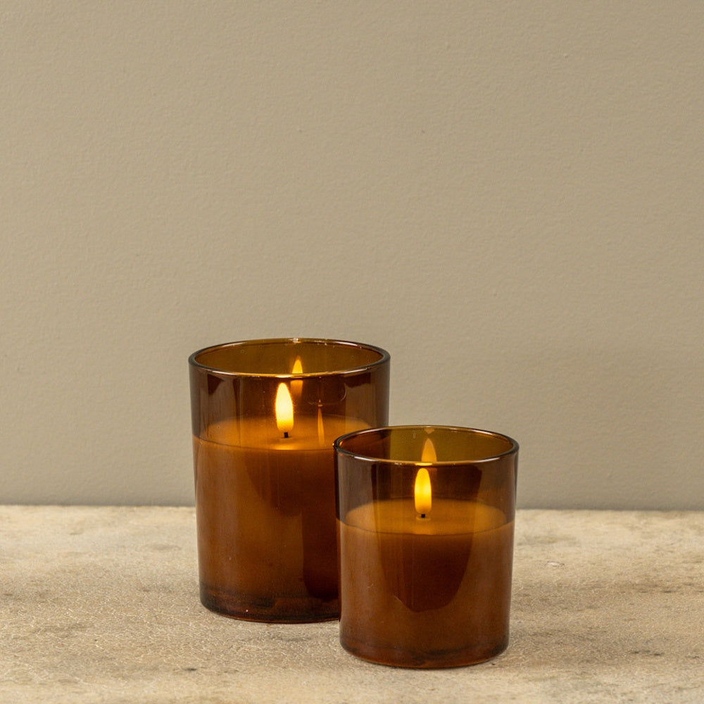 Two glowing LED candles in amber glass jars sitting on a light stone surface against a beige wall.