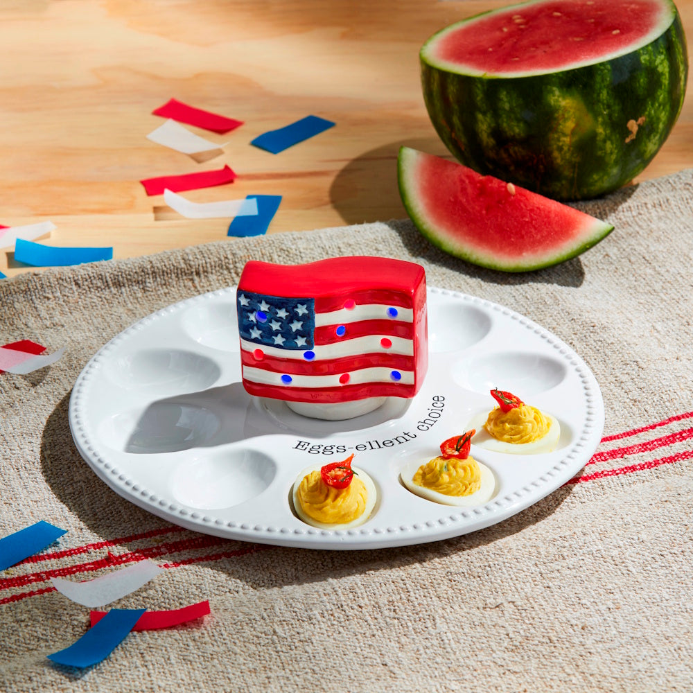 A light-up American flag sitter on a white deviled egg tray with watermelon and confetti for a patriotic celebration.