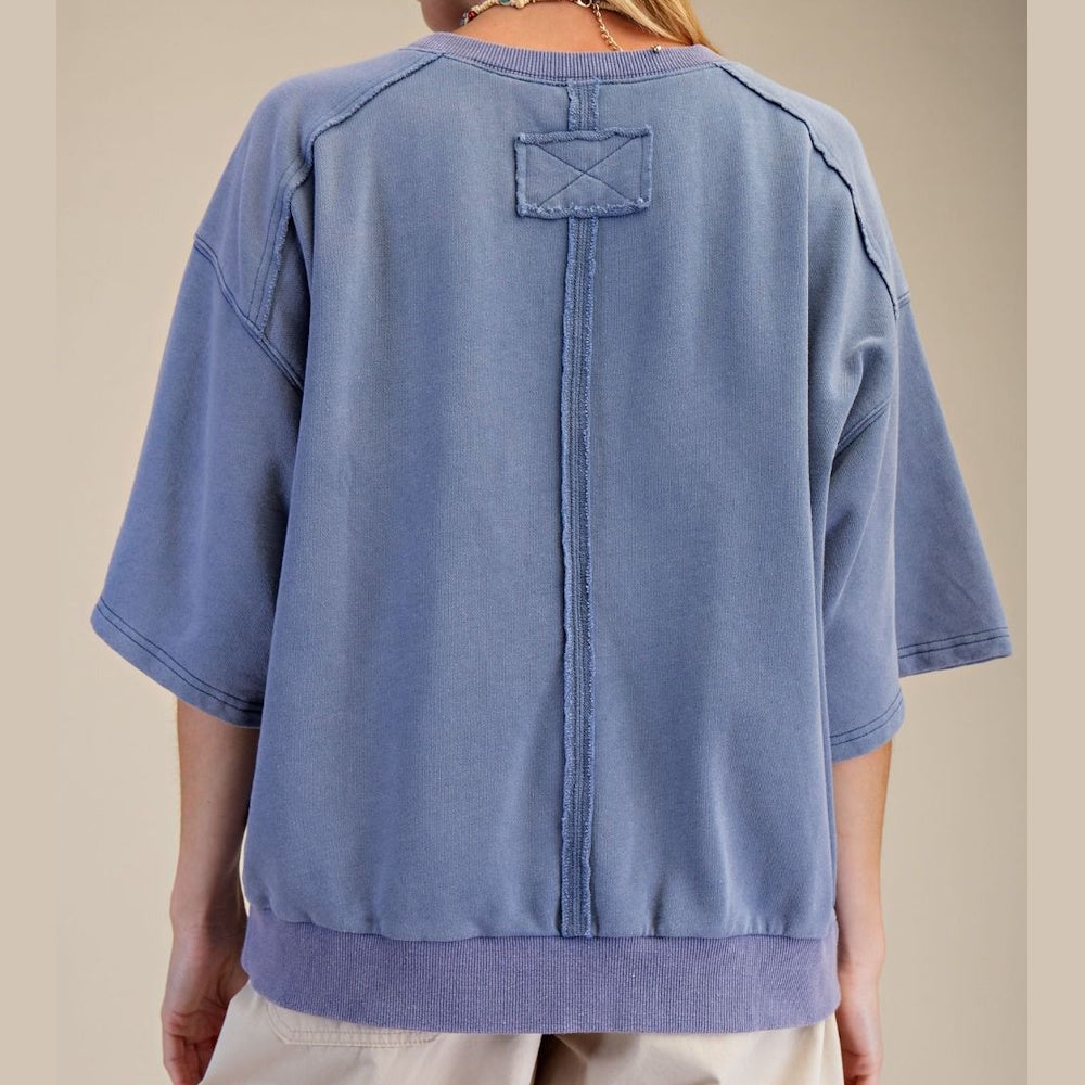 Back view of oversized blue short sleeve sweatshirt with raw seam down center and square patch detail.