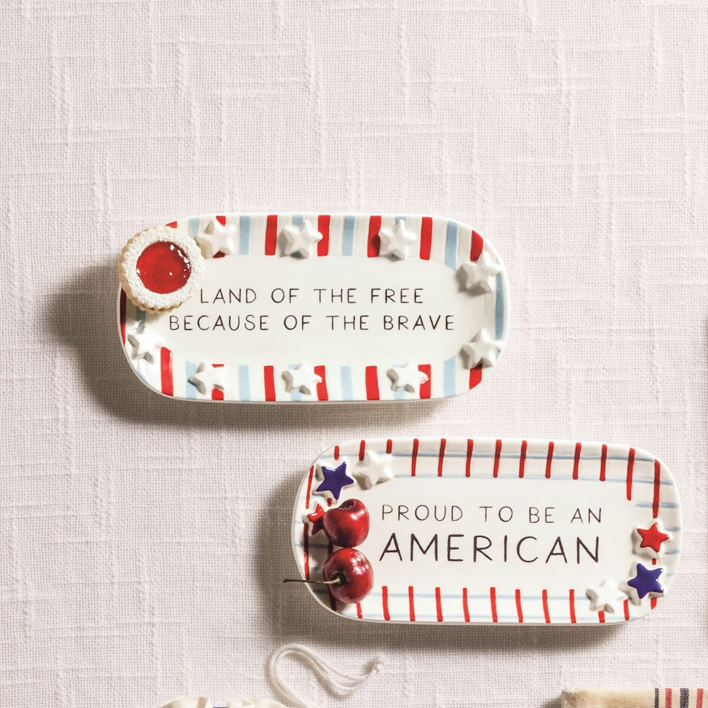 Two Americana ceramic trays with red and blue stripes, stars, and patriotic quotes on a white textured tablecloth.