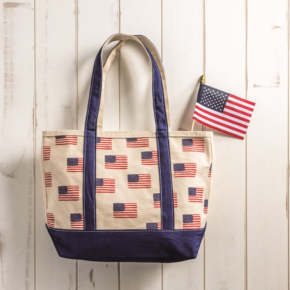 Americana canvas tote bag with an all-over US flag print and navy blue trim, hanging against a white wooden wall.