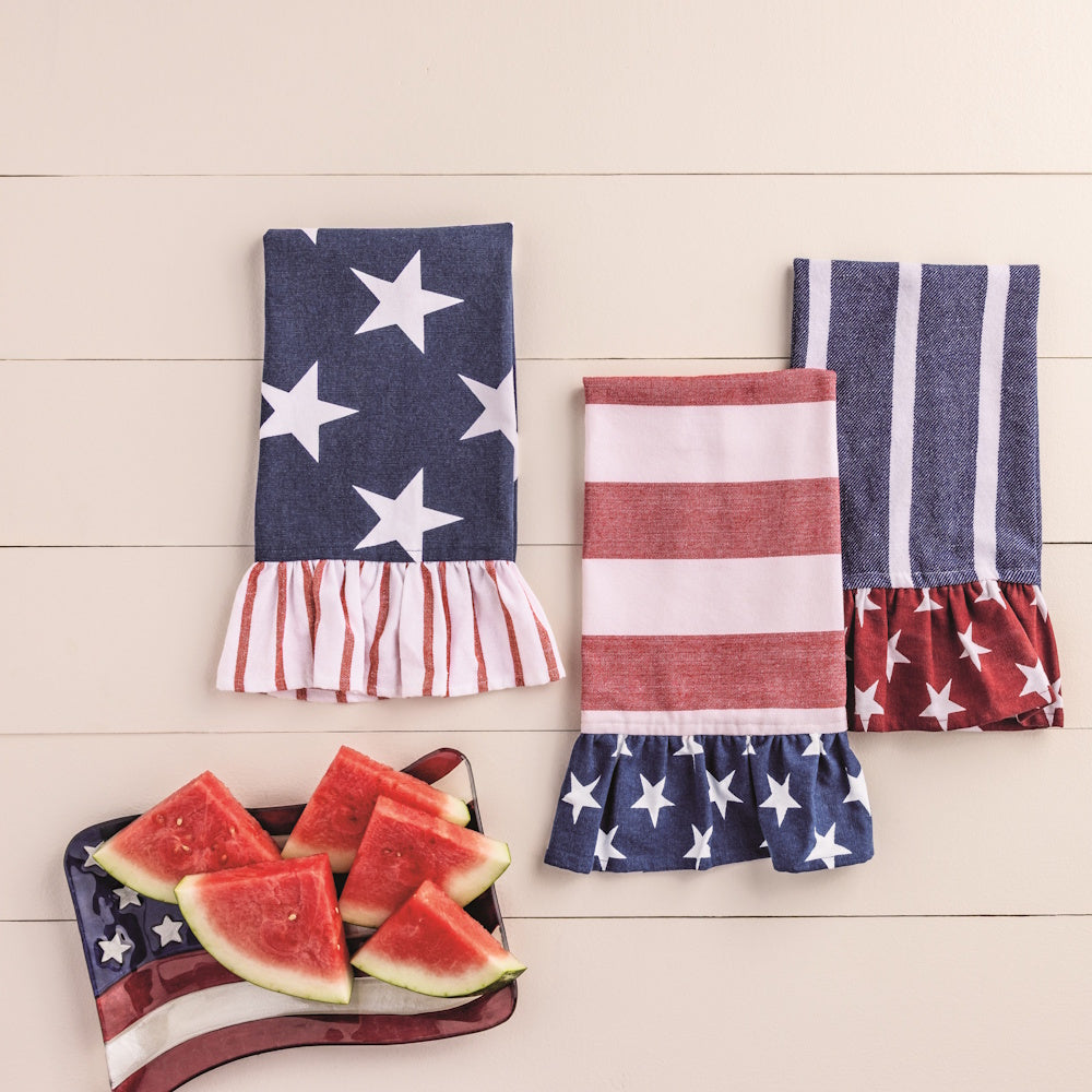 Three Americana-themed kitchen towels with ruffles and star patterns next to a plate of watermelon slices on a white table.