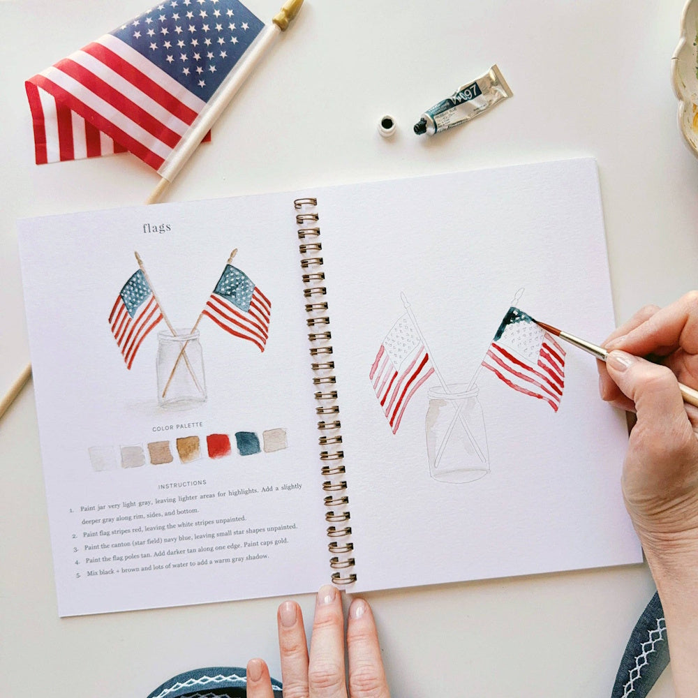 Watercolor workbook page showing American flags in a jar with painting instructions and a hand holding a brush.