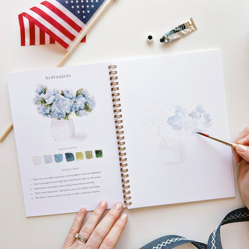 Americana watercolor workbook open to a hydrangea tutorial with paint swatches, a brush, and a US flag.