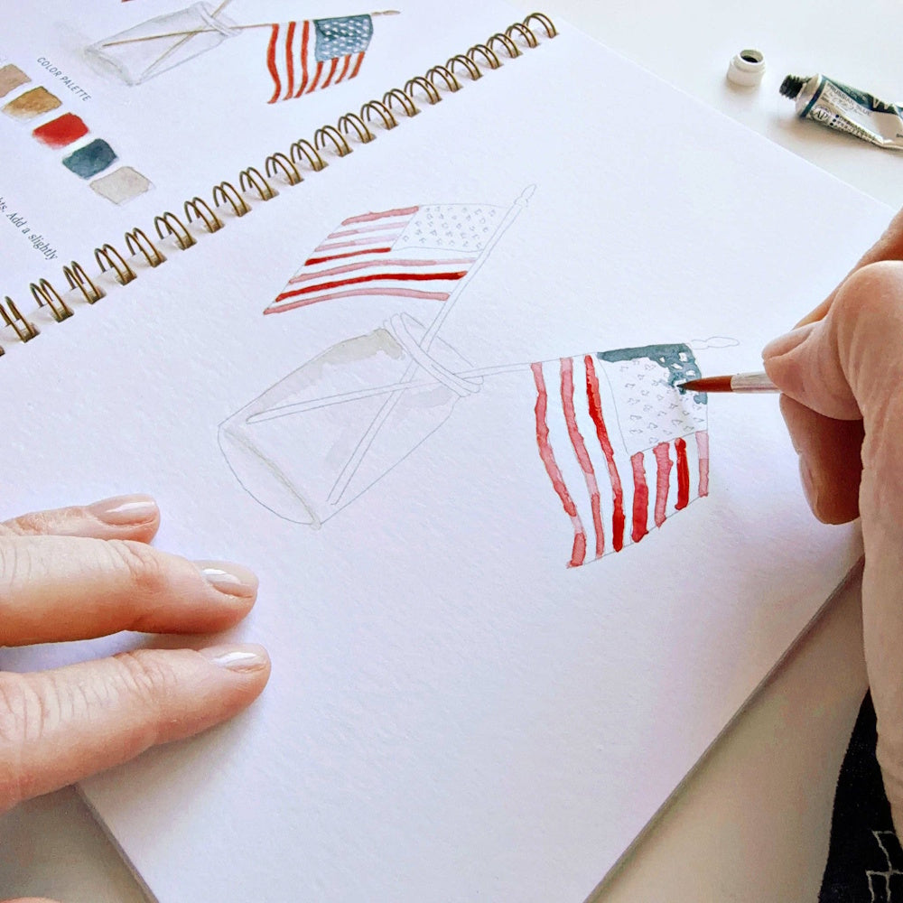 Watercolor artist painting American flags in a mason jar within a spiral workbook; simple, patriotic art style.