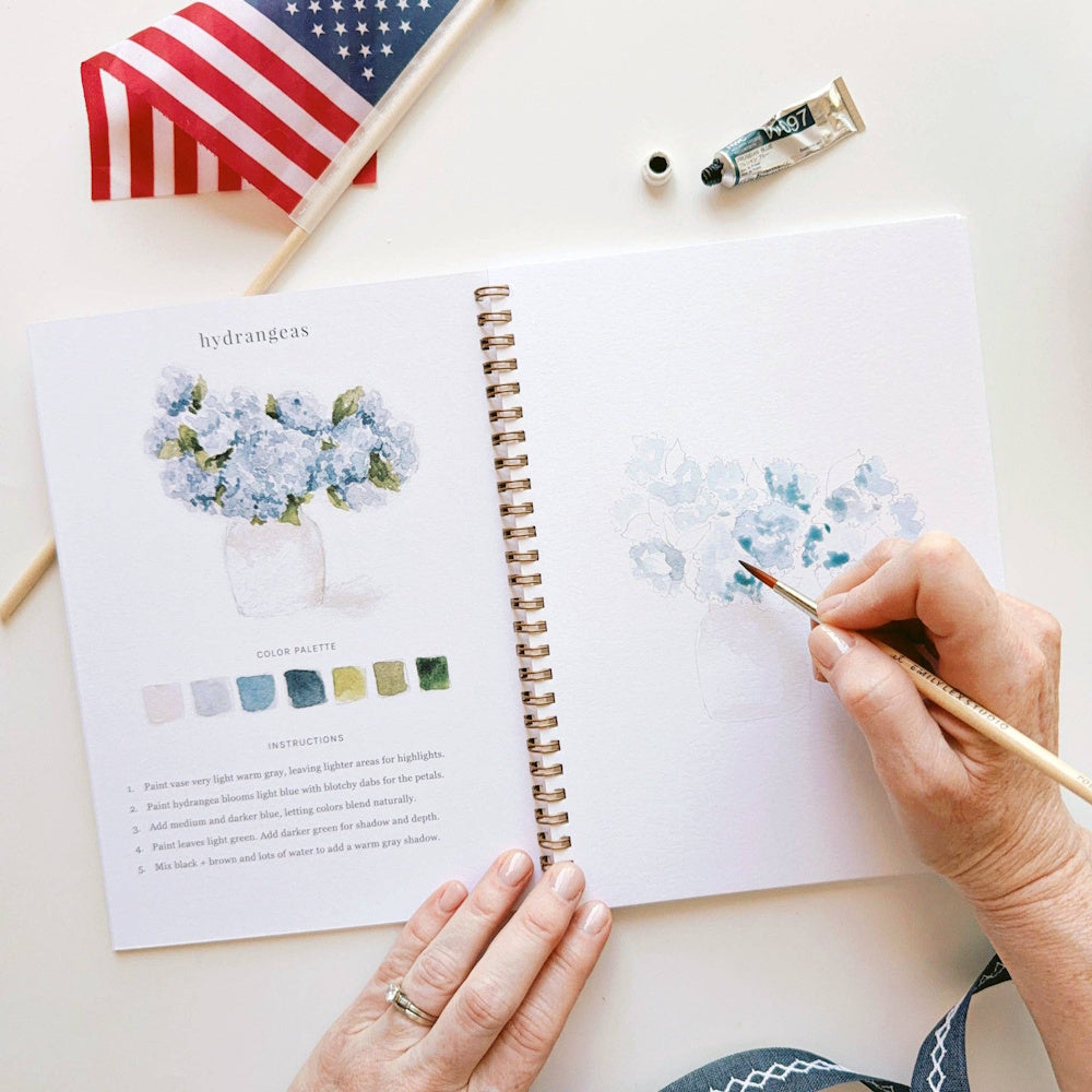 Watercolor hydrangea painting tutorial in an Americana workbook with blue paint, brushes, and a small American flag.