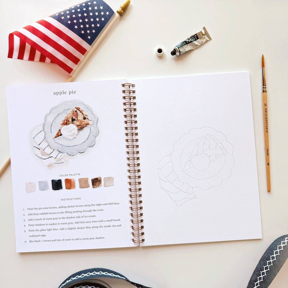 Americana watercolor workbook open to an apple pie painting tutorial with a brush, paint tube, and US flag.