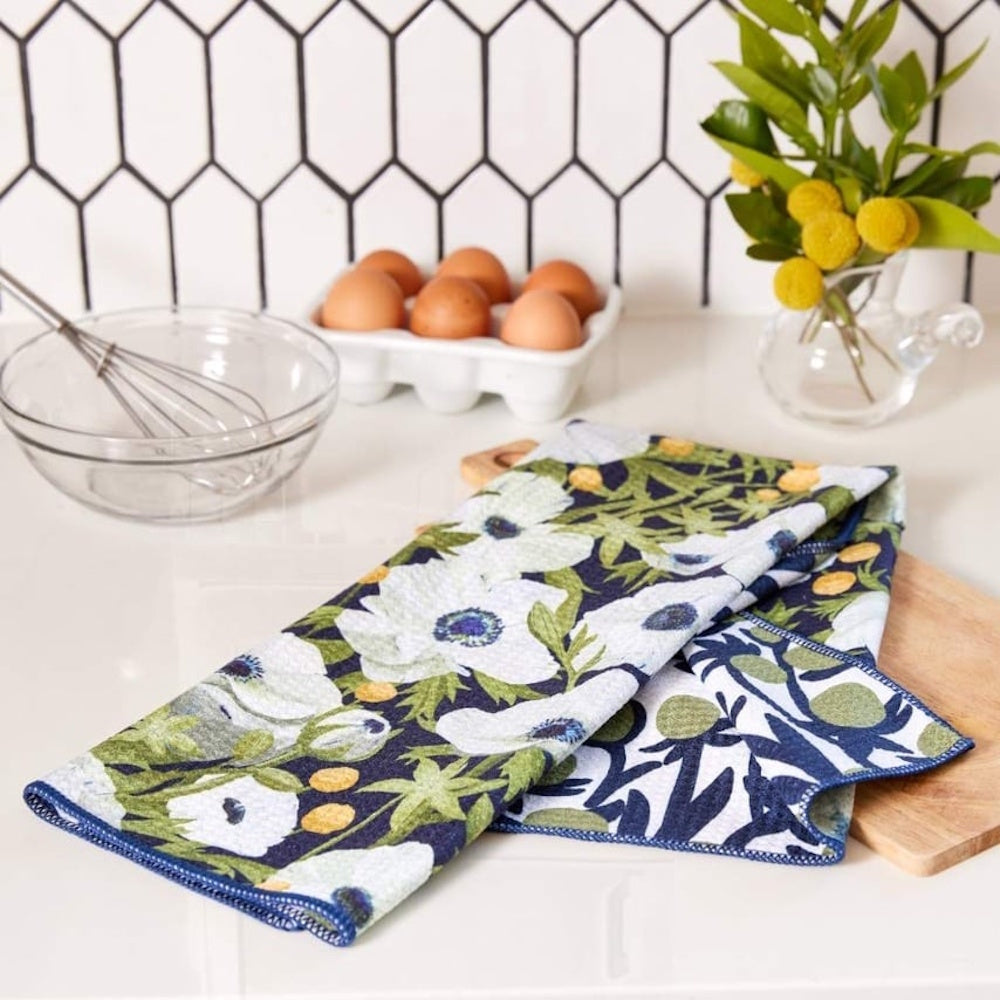Blue and white anemone poppy kitchen tea towel on a white counter with eggs, a whisk, and yellow flowers.