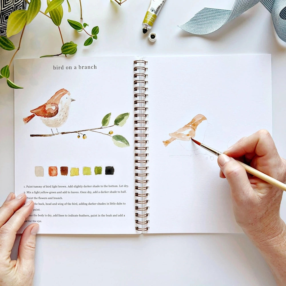 Hand using a paintbrush to color a bird illustration in an open watercolor workbook.