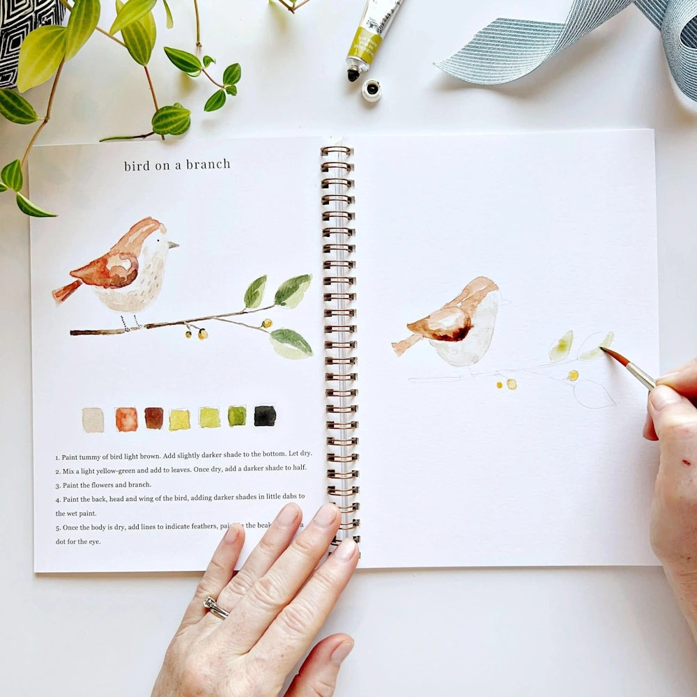 Hand painting green leaves on a bird illustration in a spiral-bound watercolor workbook.