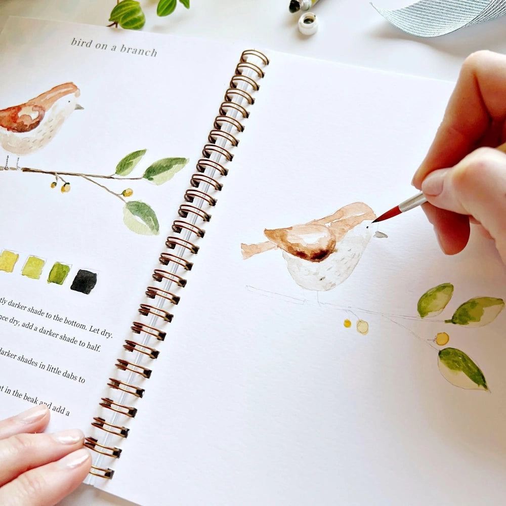 Close-up of a hand using a paintbrush to paint the eye on a watercolor bird illustration.