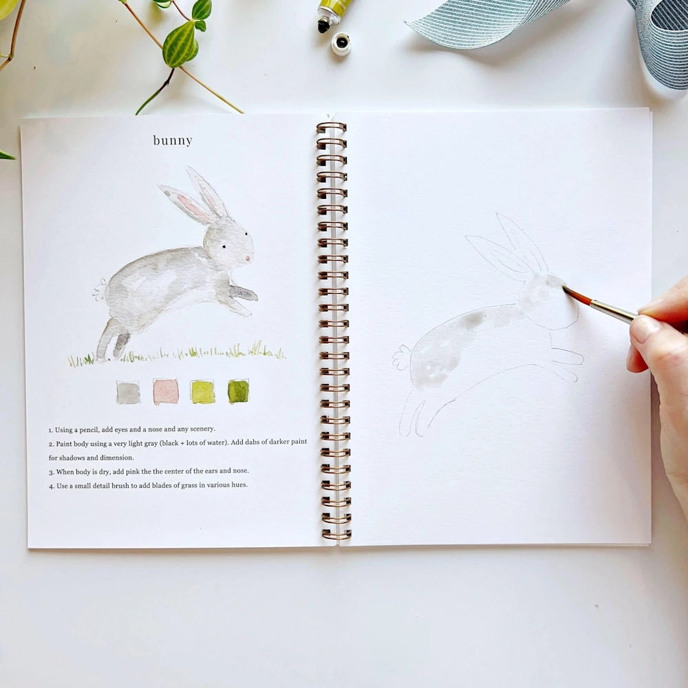Hand using a paintbrush to color a grey bunny illustration in an open watercolor workbook.