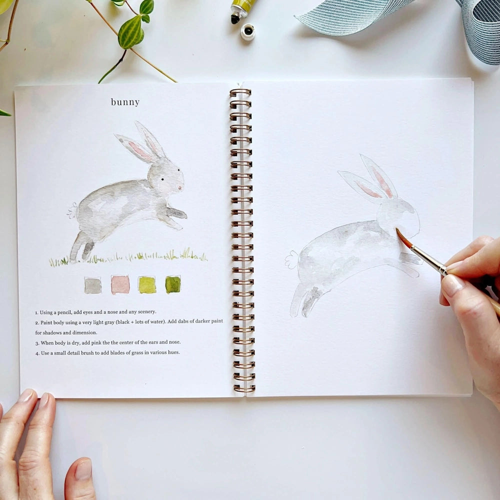 Hand adding pink watercolor details to a bunny's ears in an open workbook.