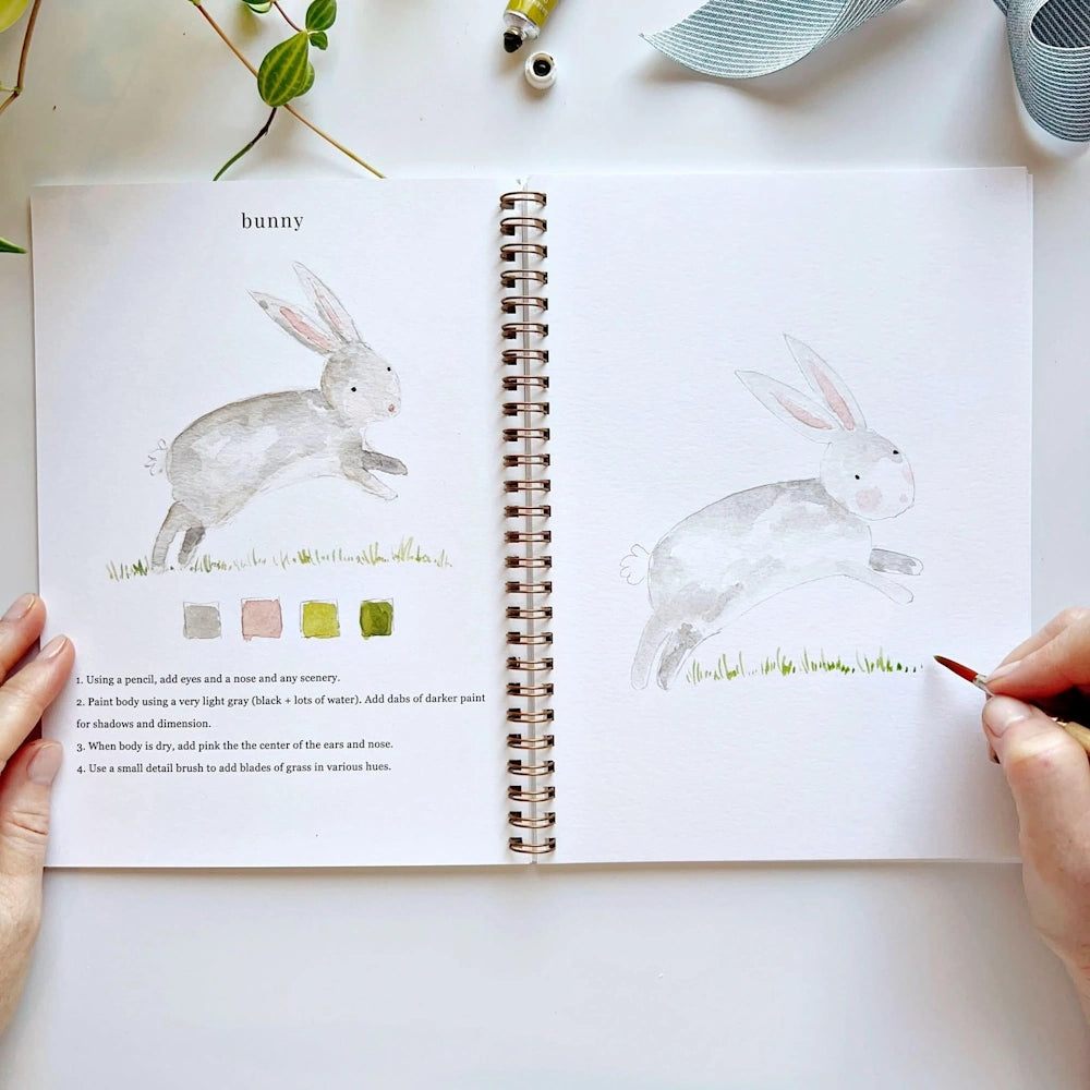 Hand painting green blades of grass beneath a watercolor bunny illustration in a workbook.