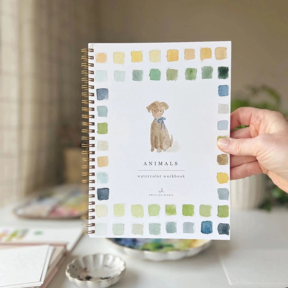 Hand holding an Animals watercolor workbook with a dog illustration and colorful paint swatches.