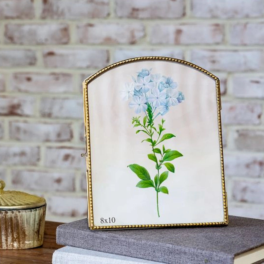Antique brass arch standing photo frame with blue floral print on a stack of books against a white brick wall.