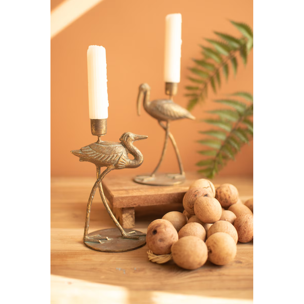 Two antique brass shorebird candleholders with white taper candles set on a tabletop decor setup.