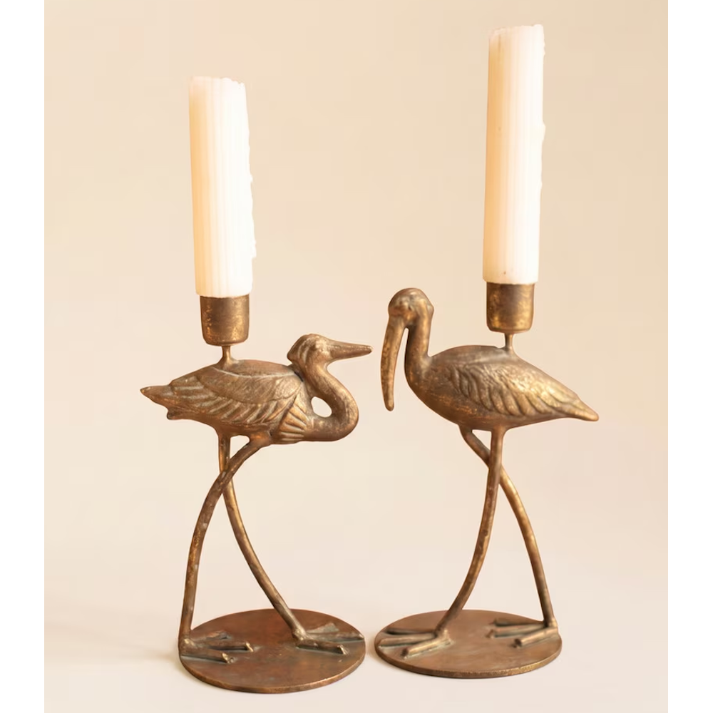 Two antique brass shorebird taper candleholders holding white candles against a plain background.