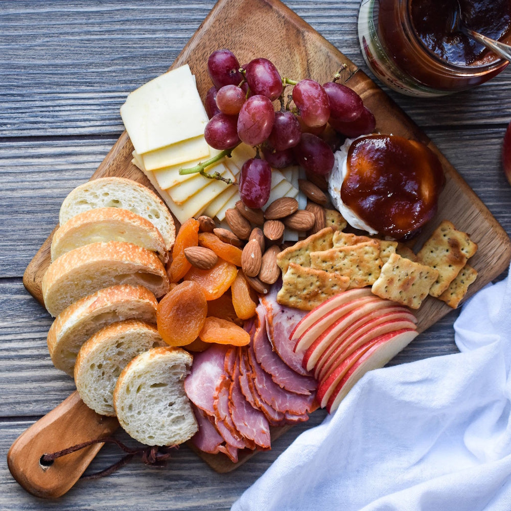 A wooden charcuterie board filled with sliced bread, meats, cheeses, grapes, apple slices, and a jar of apple butter.