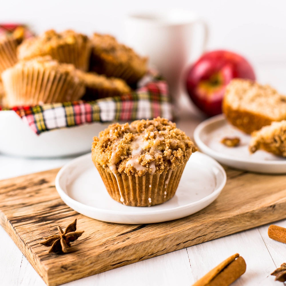 A glazed crumb muffin on a small plate and wooden board, surrounded by more muffins, a red apple, and spices.
