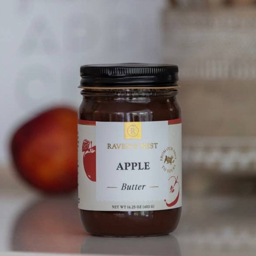 A glass jar of Raven's Nest Apple Butter with a white label, set against a blurred background with an apple.