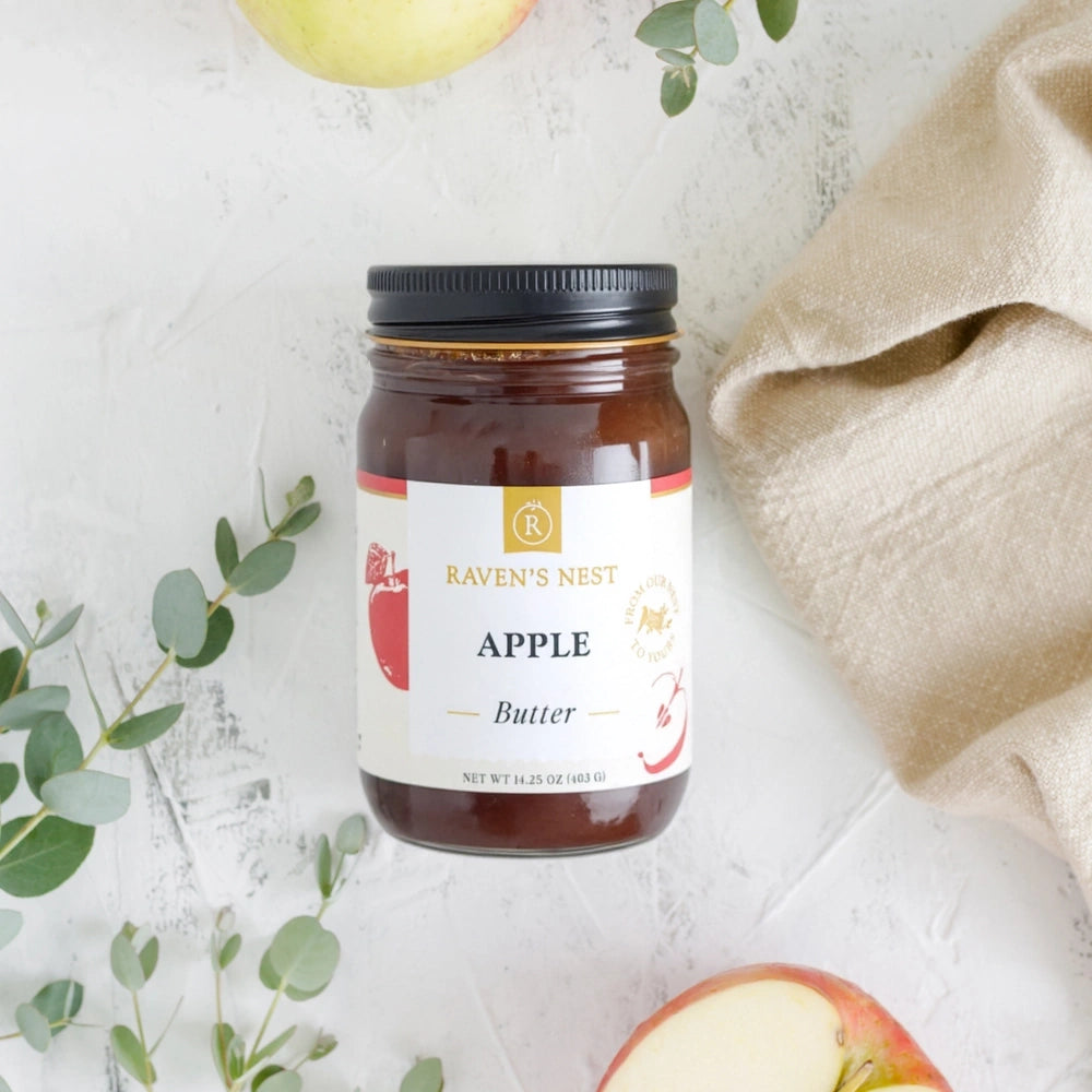 A jar of Raven's Nest Apple Butter on a white surface, surrounded by fresh apples and green leaves.